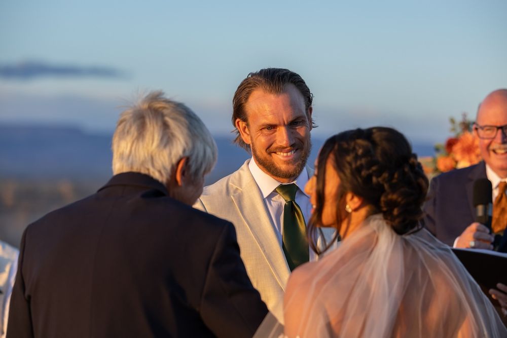 Groom in white suit smiling at wedding guests during outdoor ceremony with mountain views at golden hour.