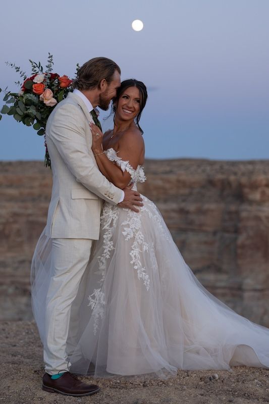 Bride and groom embrace at canyon edge at dusk with full moon, she holds floral bouquet in lace gown.