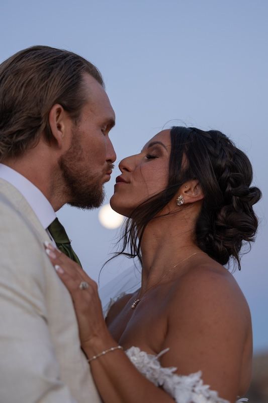 Bride and groom share a romantic kiss at dusk with the moon glowing softly behind them on their wedding day.