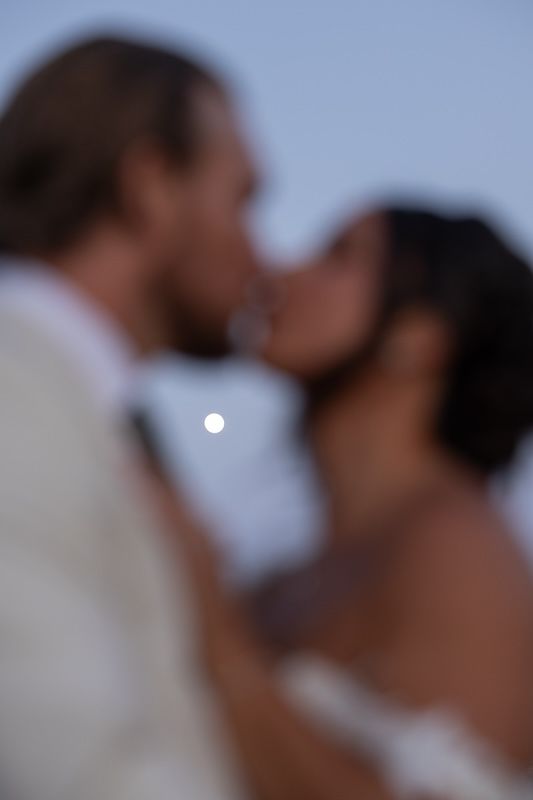Blurred couple sharing a romantic kiss with the moon visible between them at dusk.