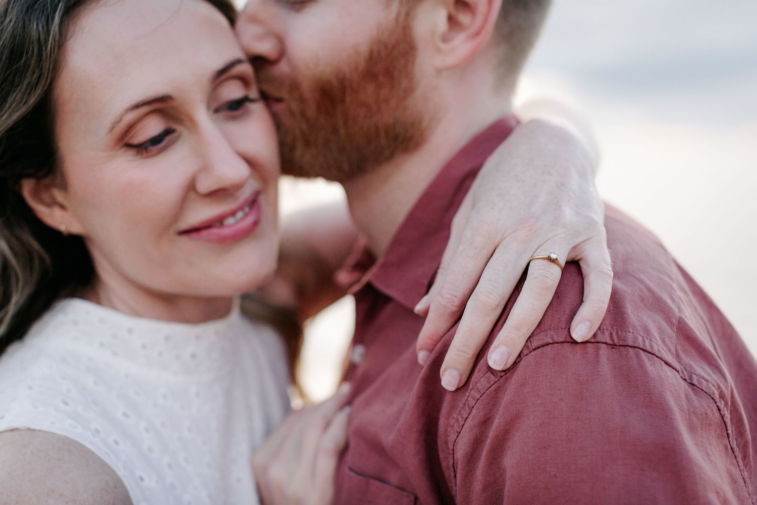 A couple shares a tender moment, the woman smiling as her partner kisses her cheek, showcasing an engagement ring.