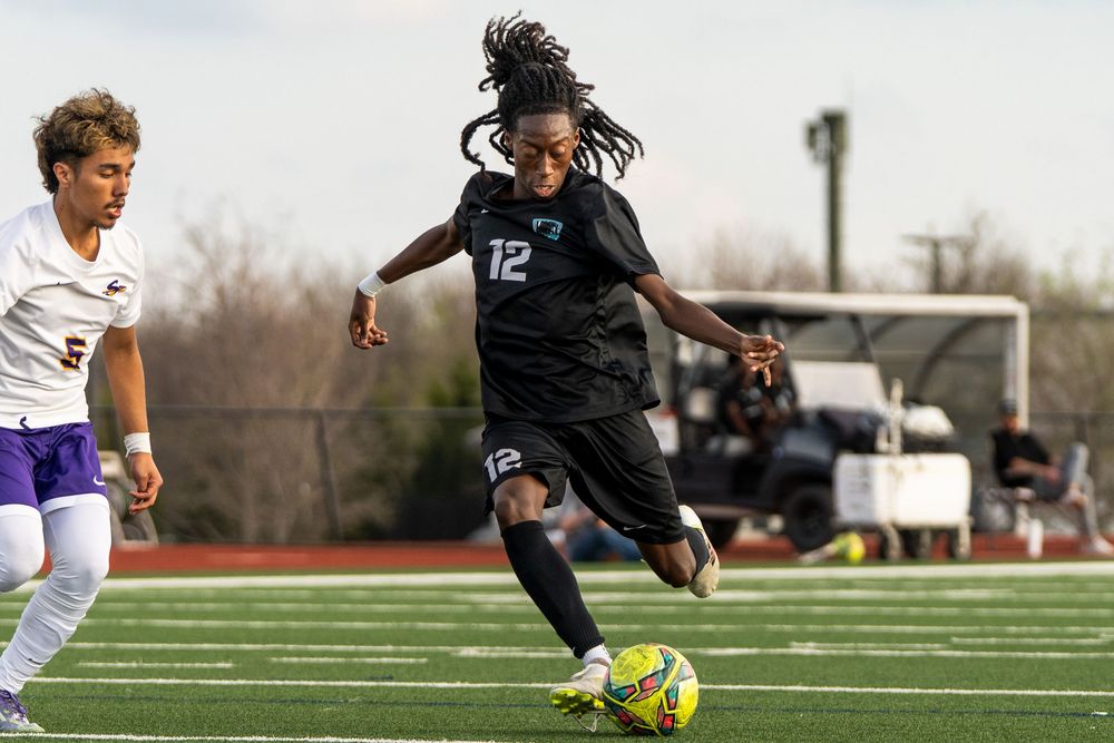 Soccer player number 12 in black uniform dribbling ball during competitive match against purple-uniformed opponent.