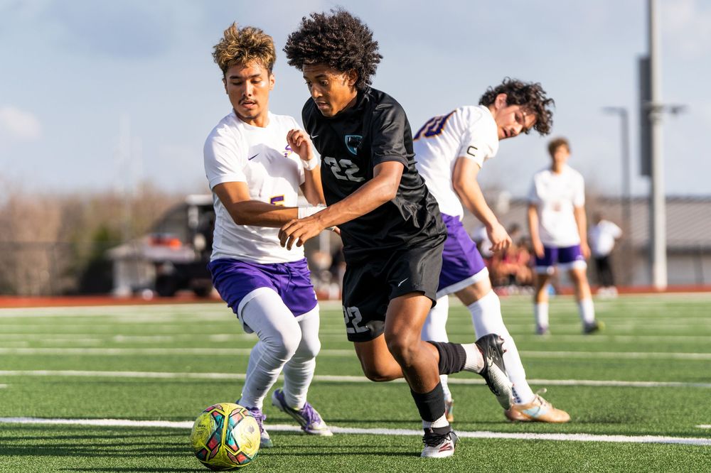Soccer player number 22 in black uniform competing for ball against two purple-uniformed defenders on field.