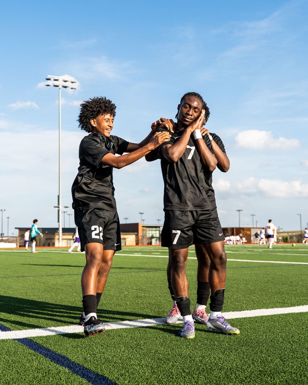 Two soccer players in black uniforms celebrating together on green field under blue sky after scoring.