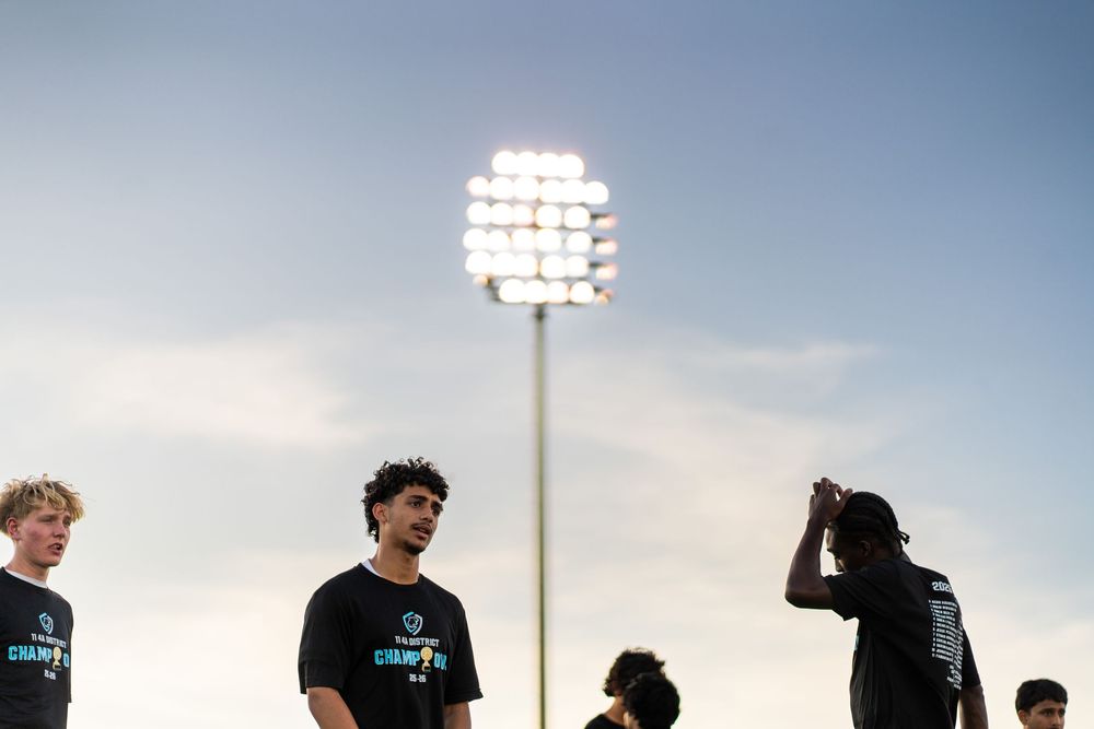 Soccer players standing on field under bright stadium lights at dusk.