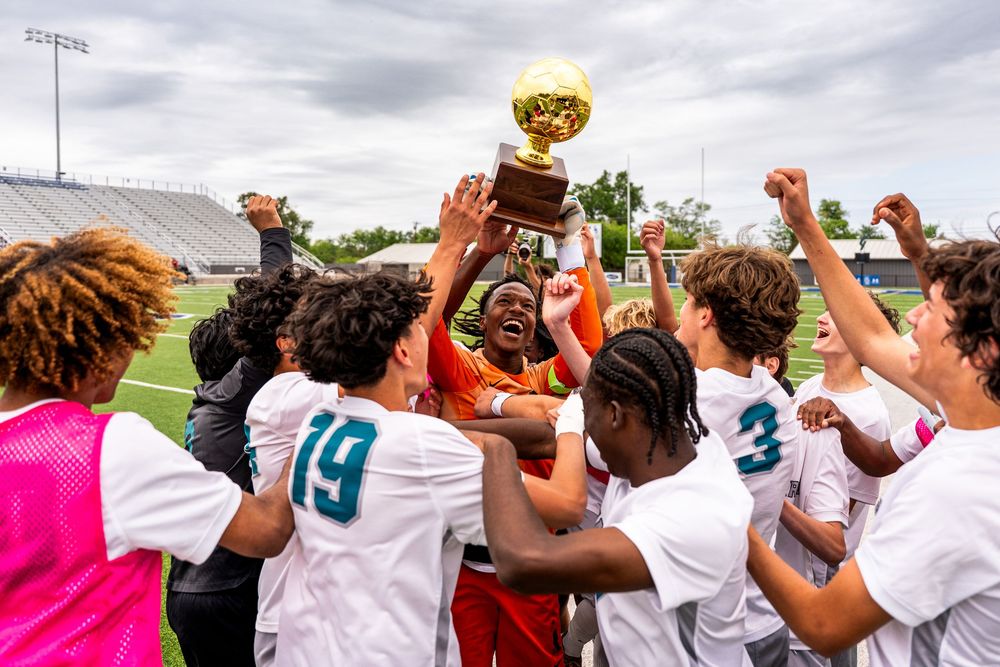 High school soccer team celebrating championship win by lifting golden trophy together on field.