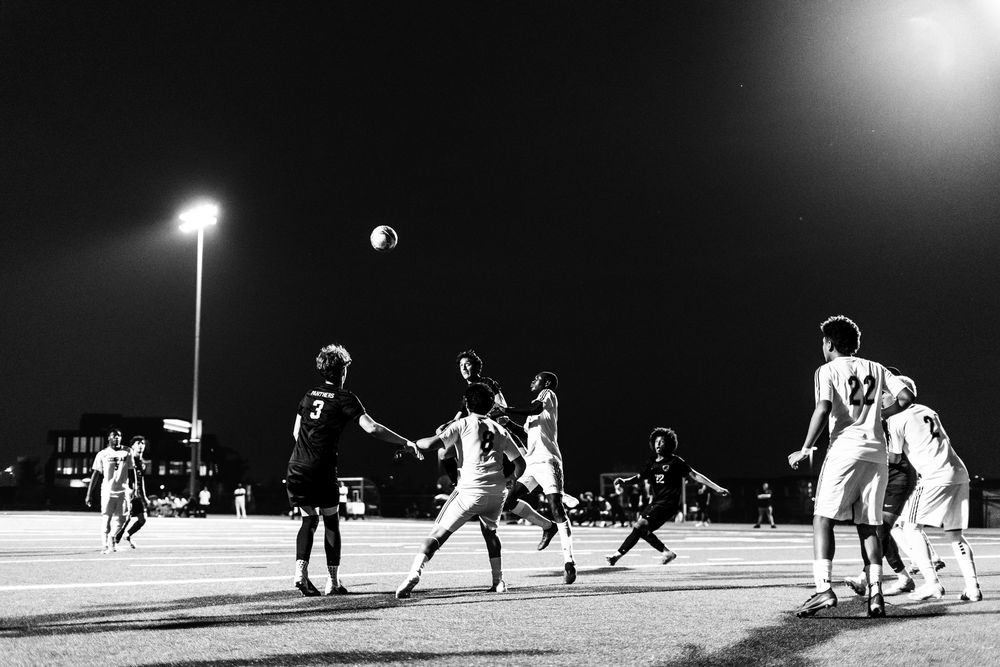 Black and white photo of soccer players competing for header during floodlit night match.