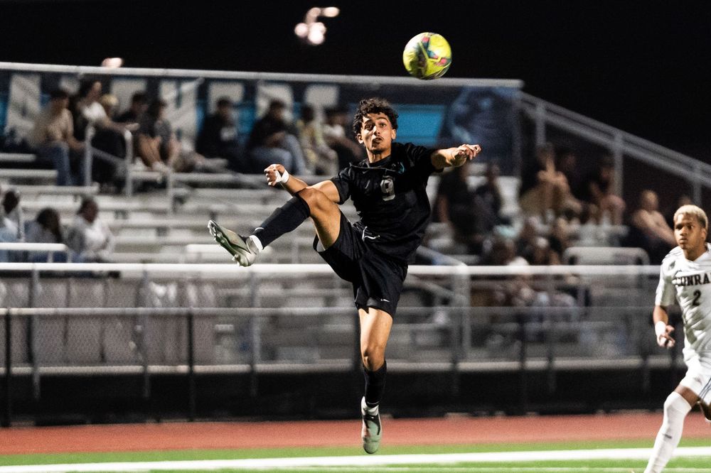 Soccer player in black uniform performing aerial kick during night game on stadium field.