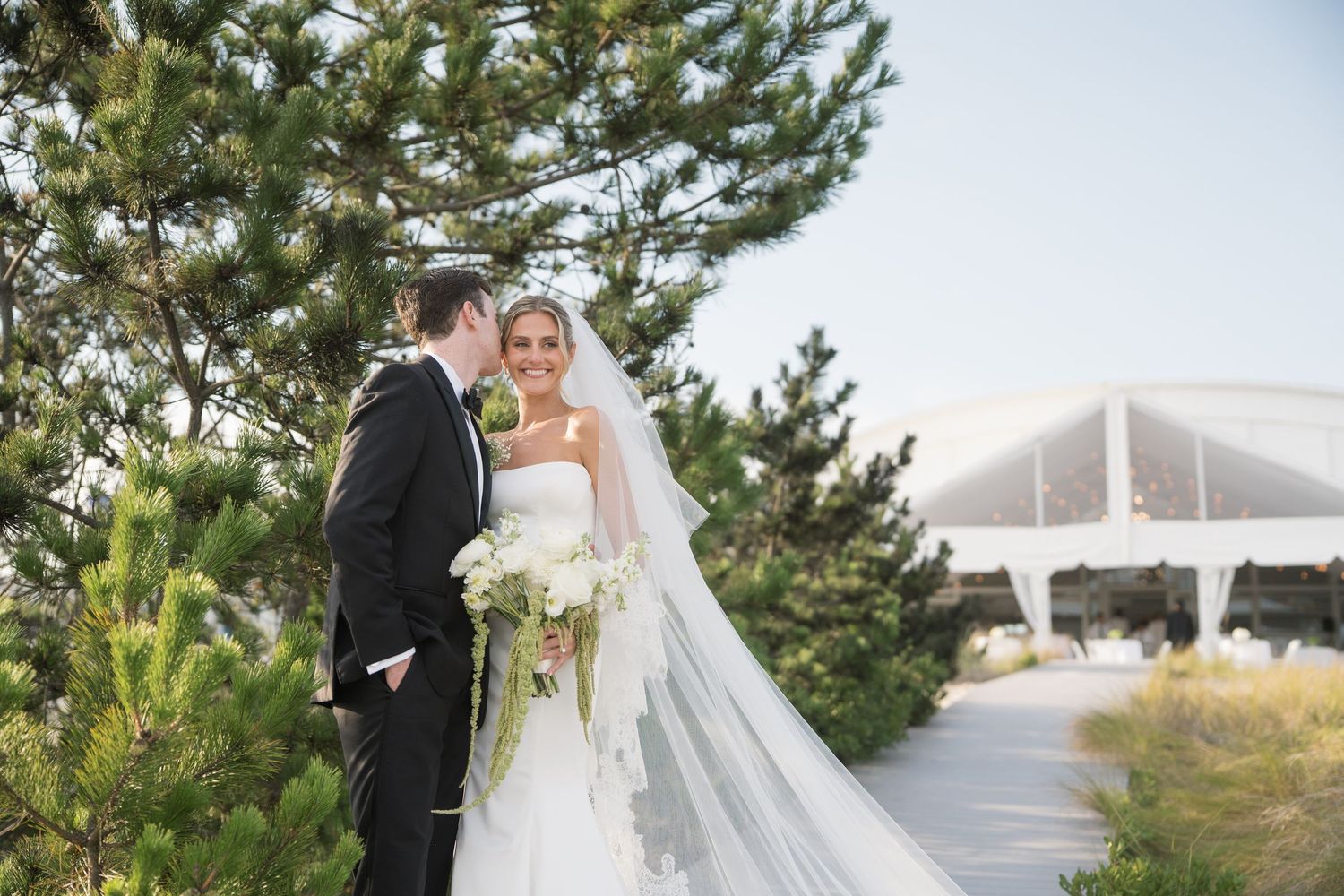 A flowing white wedding dress train trails behind a couple sitting on stone steps during an outdoor photoshoot.