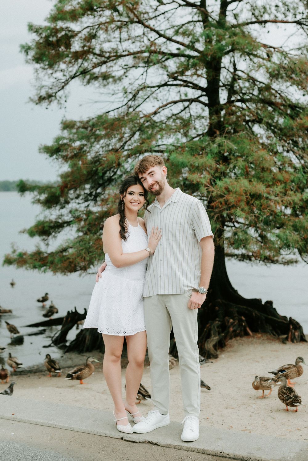 Couple posing by lakeside with ducks and large tree, woman in white dress, man in striped shirt and khakis.