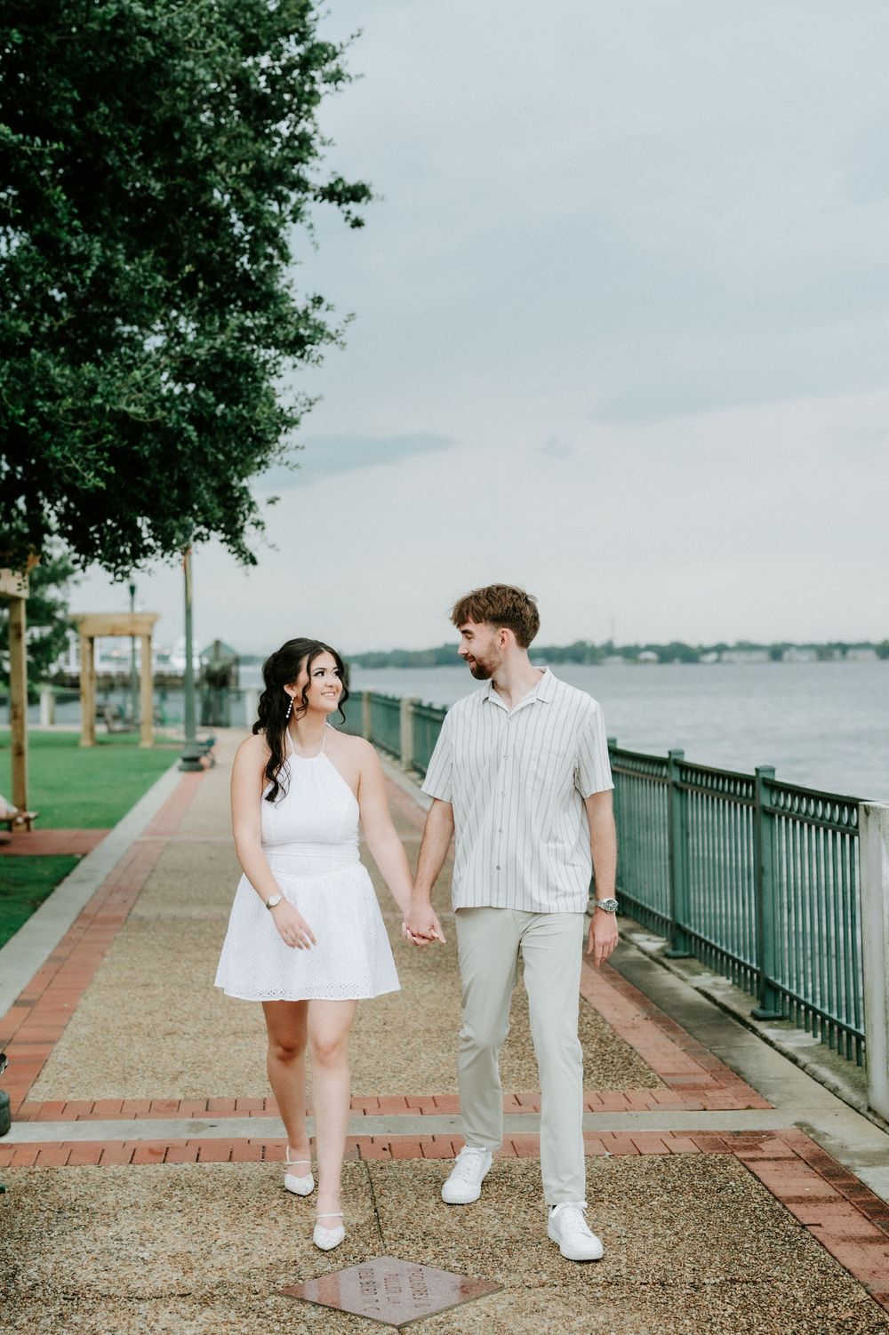 Couple holding hands walking along a waterfront boardwalk, woman in white dress, man in striped shirt and khakis.
