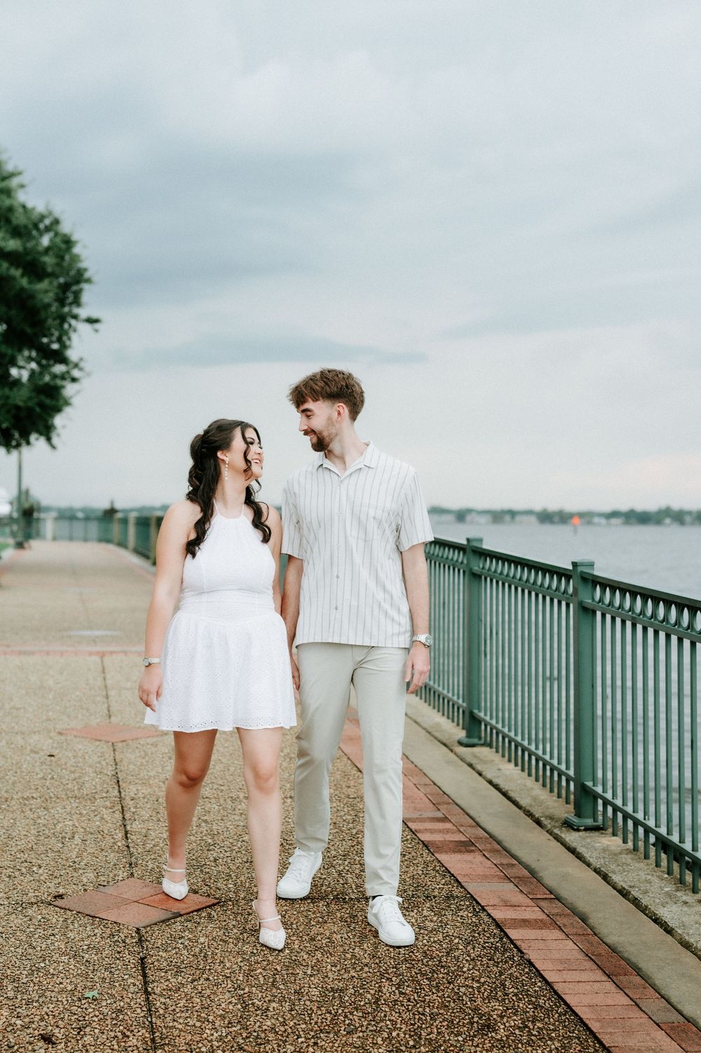 A couple holds hands and smiles at each other on a waterfront promenade, woman in white dress, man in striped shirt.