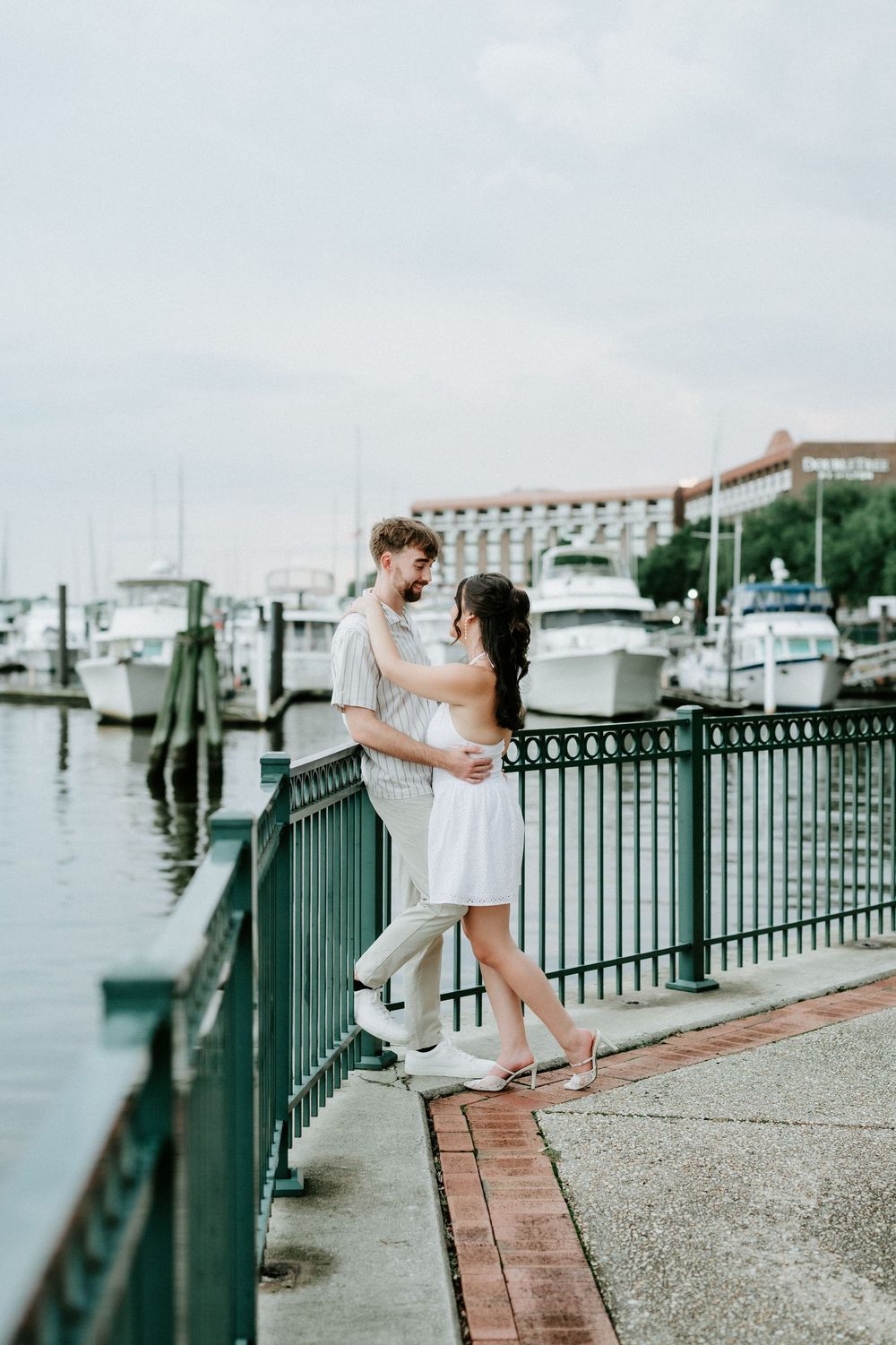 Couple embracing on a waterfront dock with marina boats and a hotel in the background on a cloudy day.