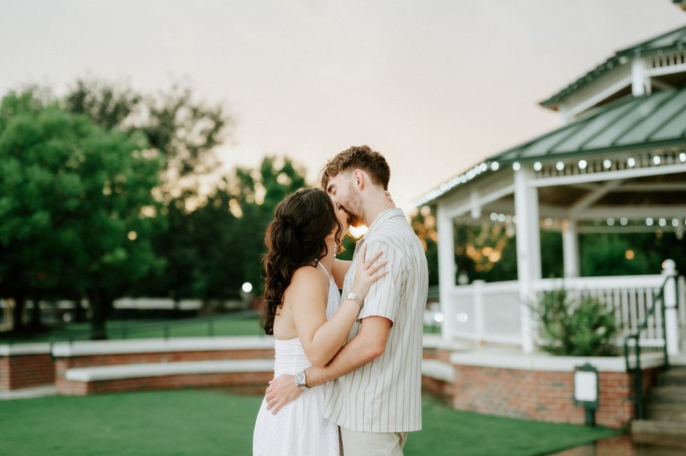 Couple sharing a romantic kiss outdoors near a white gazebo at sunset, dressed in elegant white outfits.