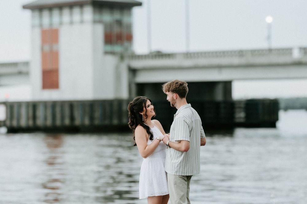 Couple holding hands and smiling at each other by the waterfront with a bridge in the background.