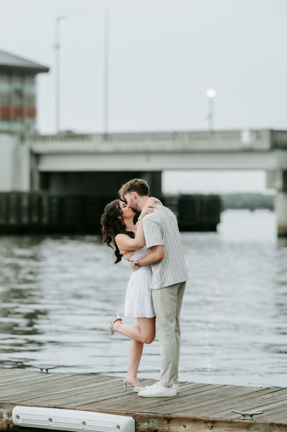 Couple sharing a romantic kiss on a waterfront dock, woman in white dress, man in striped shirt, bridge in background.