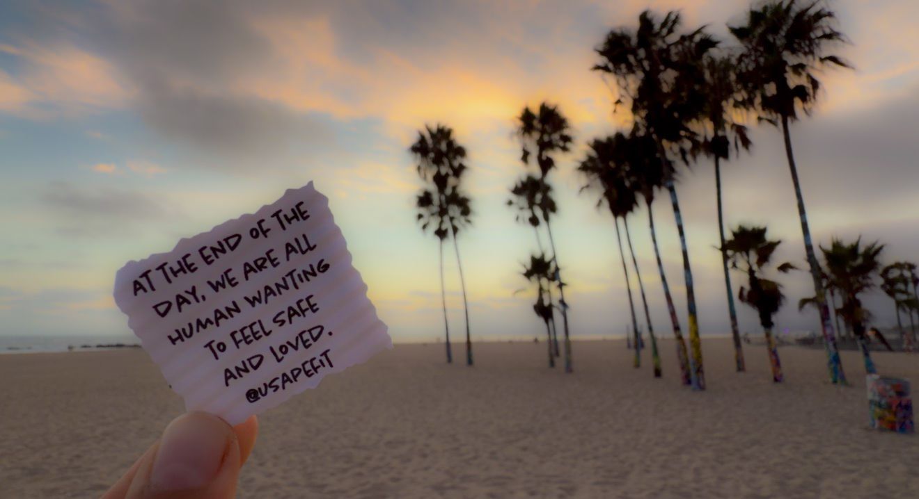 Hand holding a note on Venice Beach at sunset with palm trees, reading inspirational quote about feeling safe and loved.