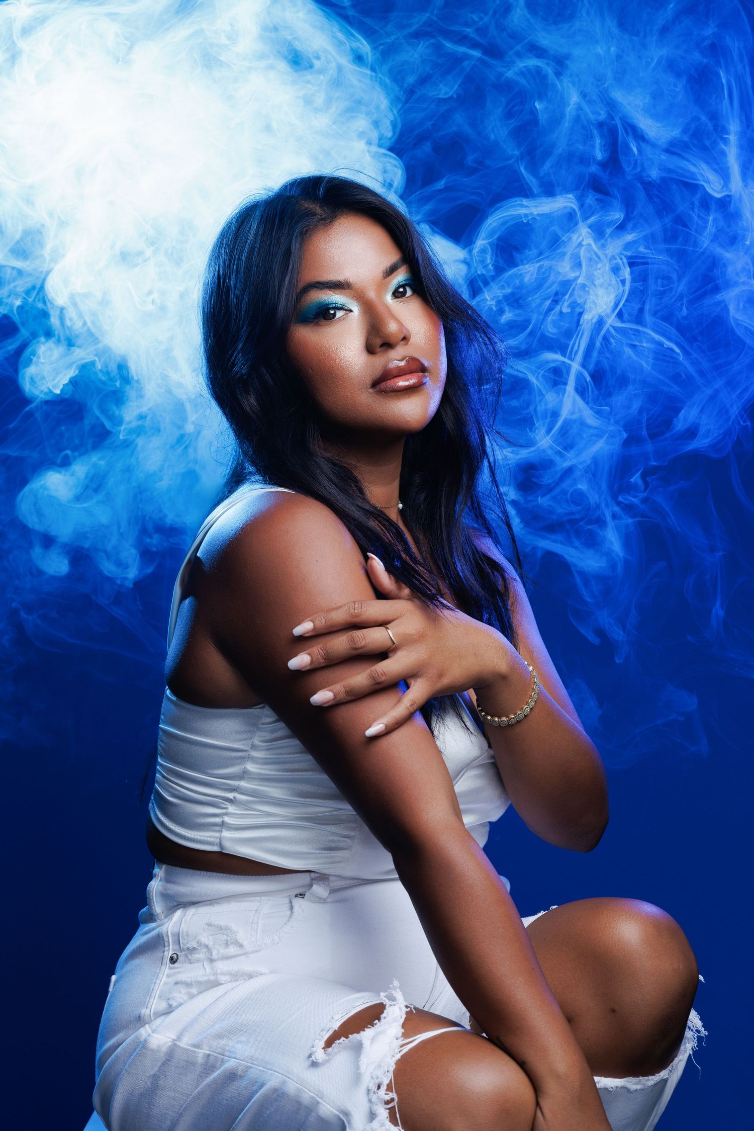 A person in a white t-shirt and distressed jeans sits barefoot against a light studio background, showing red nail polish.