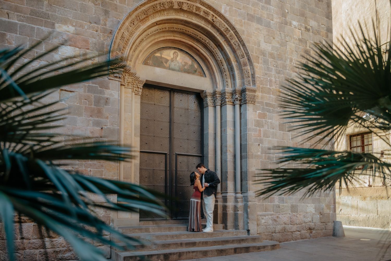A smiling engaged couple walking hand-in-hand down a narrow, sunlit cobblestone street in the historic Gothic Quarter of Barcelona.