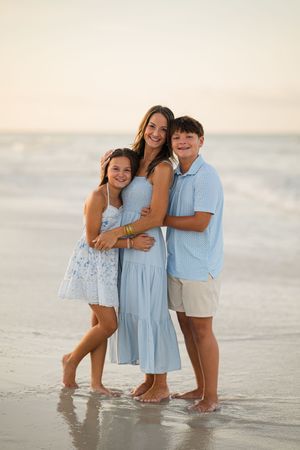 A mother and two children pose together on a sandy beach in matching blue outfits with ocean waves behind them.