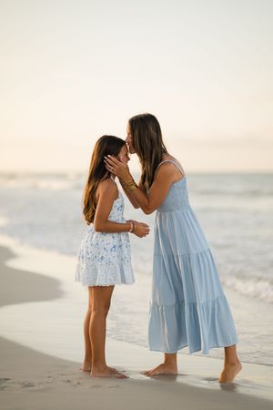 A mother and daughter share a tender moment on the beach, both wearing light blue dresses at sunset.