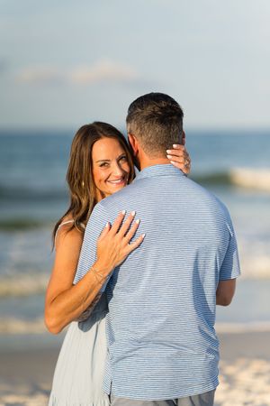 A smiling woman embraces a man in a striped shirt on a sunny beach, showcasing an engagement ring.