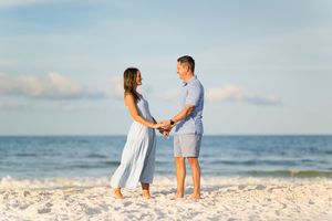Couple holding hands on a white sandy beach with ocean backdrop, wearing casual blue outfits on a sunny day.