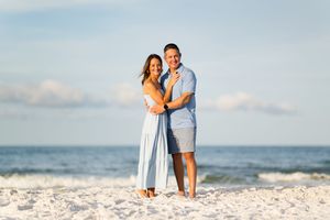 Smiling couple embracing on a white sand beach with ocean waves and blue sky in the background.