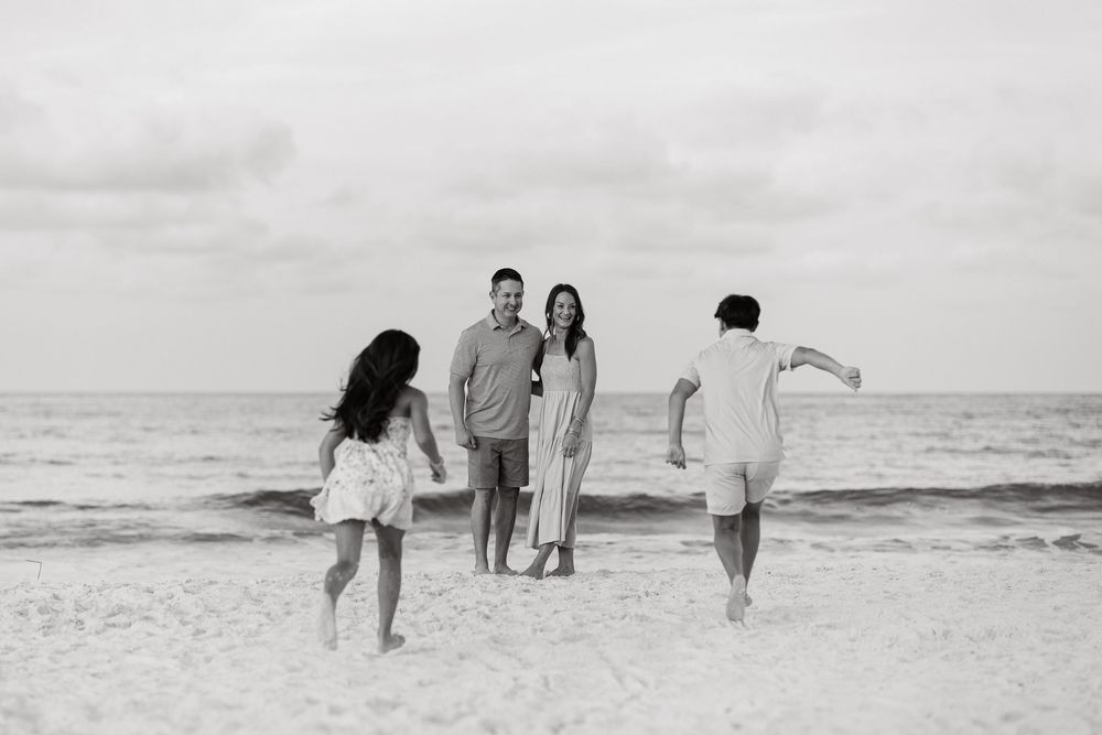 Black and white beach photo of a couple standing together as two children run toward them on a sandy shore.