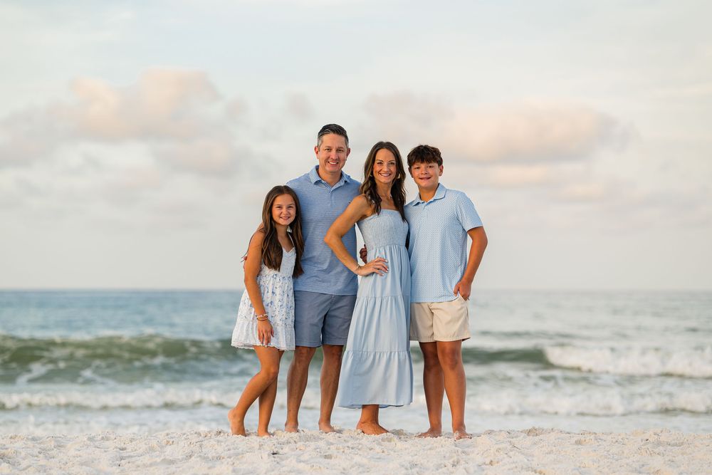 Family of four smiling together on a sandy beach with ocean waves and pastel sky backdrop.