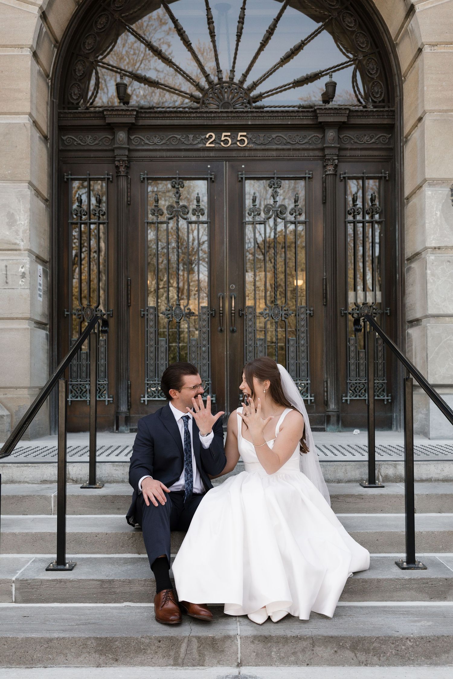 A bride and groom hold hands walking up stone steps outside a grand historic building on their wedding day.