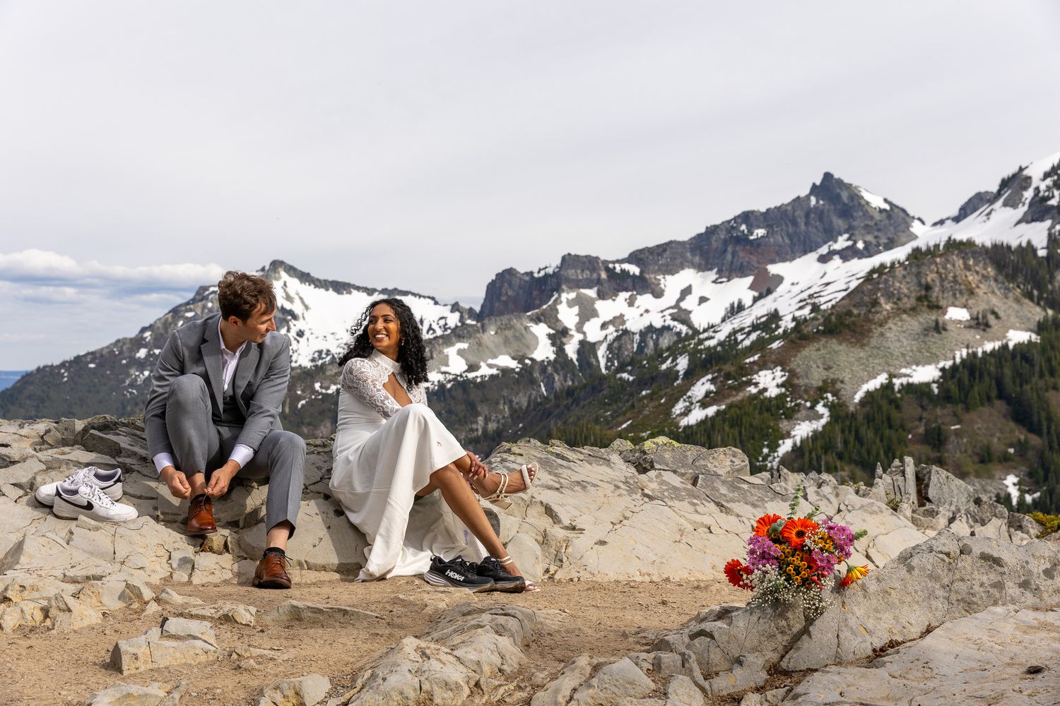 Bride and groom sitting on rocky terrain with dramatic snow-capped mountains behind them. PNW elopement photographer.