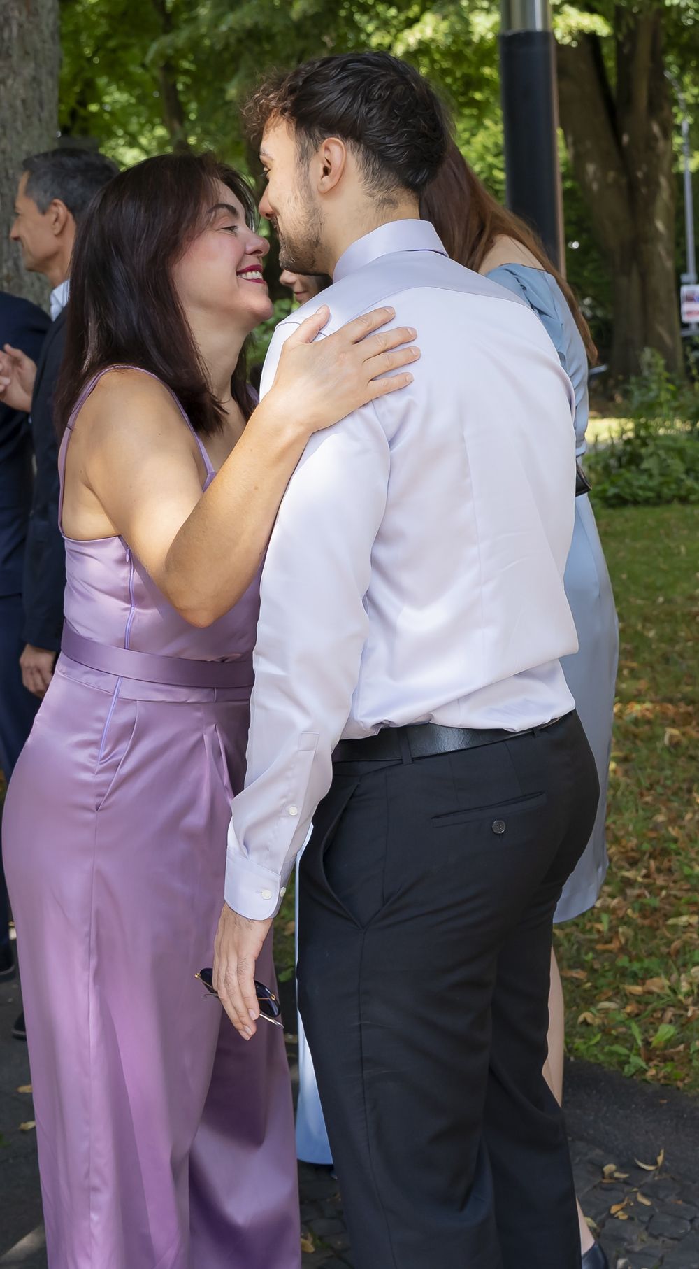 Ein Paar teilt einen intimen Moment bei einer Veranstaltung im Freien, wobei die Frau ein lavendelfarbenes Seidenkleid trägt.