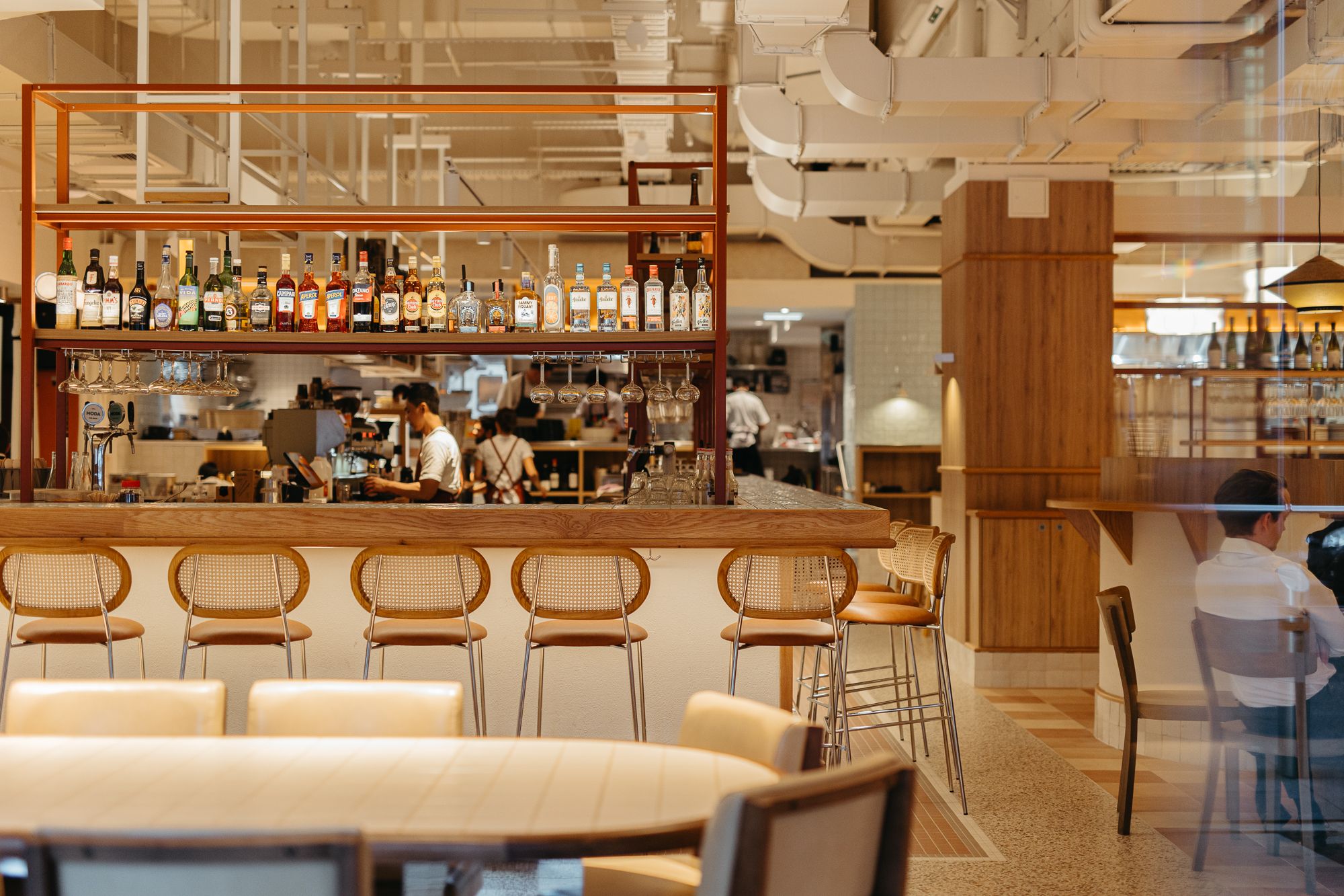 Modern restaurant bar with rattan stools, wood shelving stocked with bottles, and open kitchen in warm neutral tones.
