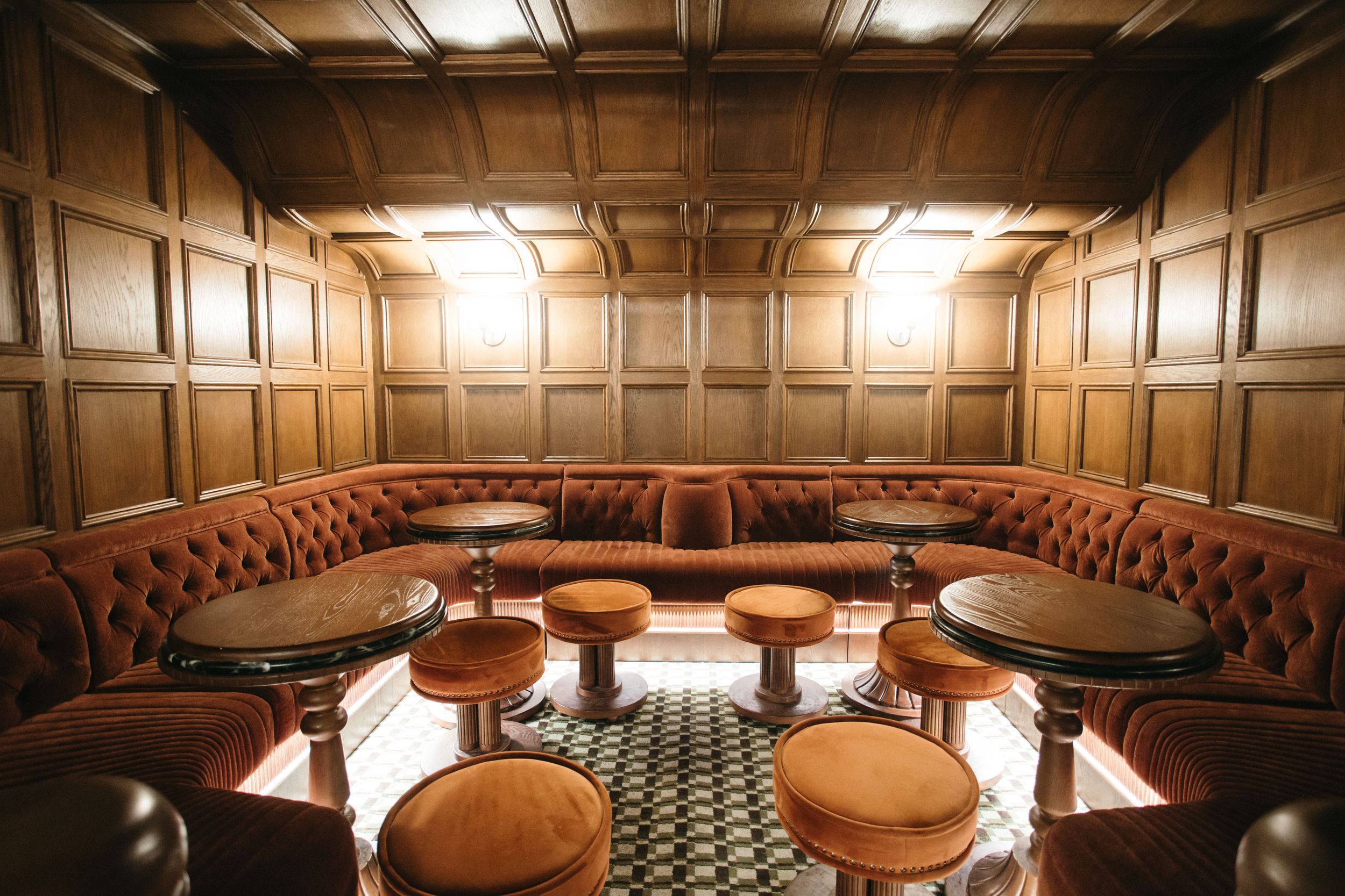 Luxurious bar interior featuring curved tufted velvet seating, wood-paneled walls, and patterned tile flooring.