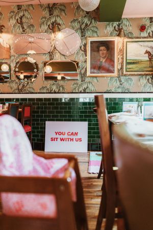 Restaurant interior with green subway tile, hanging plants, and a sign reading 'You can sit with us'.