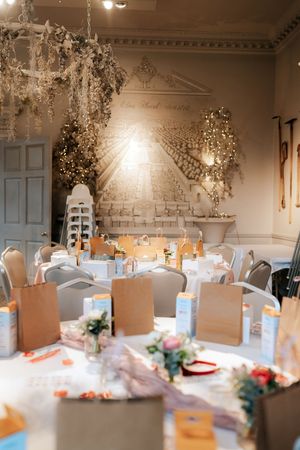 Elegant wedding reception room with round tables, floral decor, kraft paper bags, and a mural backdrop with fairy lights.