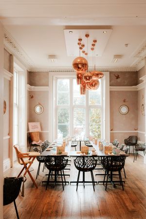 Elegant dining room with copper pendant lights, black chairs, wooden floors, and tall windows in a chic restaurant.