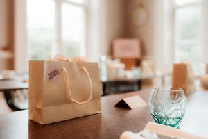 Elegant gift bag with pink ribbon bow on wooden table beside green crystal glass in bright room.
