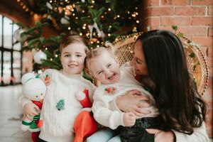 A mother with two young girls smiling near a decorated Christmas tree, holiday portrait session.