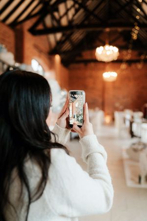Dark-haired woman in white sweater photographing festive barn venue interior with smartphone.