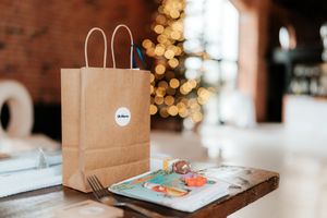 Brown kraft paper gift bag with white sticker beside colorful card on wooden board, Christmas tree bokeh background.