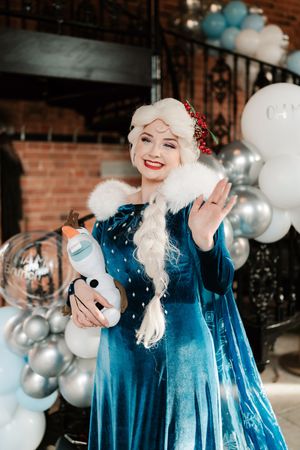 Woman dressed as Elsa from Frozen holding Olaf toy, waving at party with blue and silver balloons.