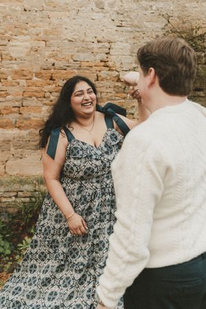 A woman in a floral dress laughs joyfully with a man in a cream sweater against a rustic stone wall.