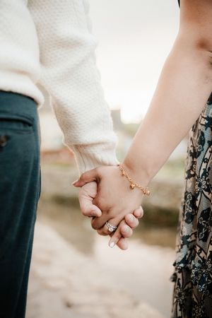 Close-up of a couple holding hands, woman wearing a diamond ring and gold bracelet, man in white sweater and dark pants.