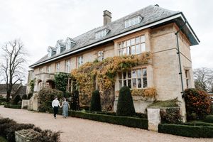 Couple walking outside a grand English manor house with stone walls and ivy, surrounded by manicured hedges.
