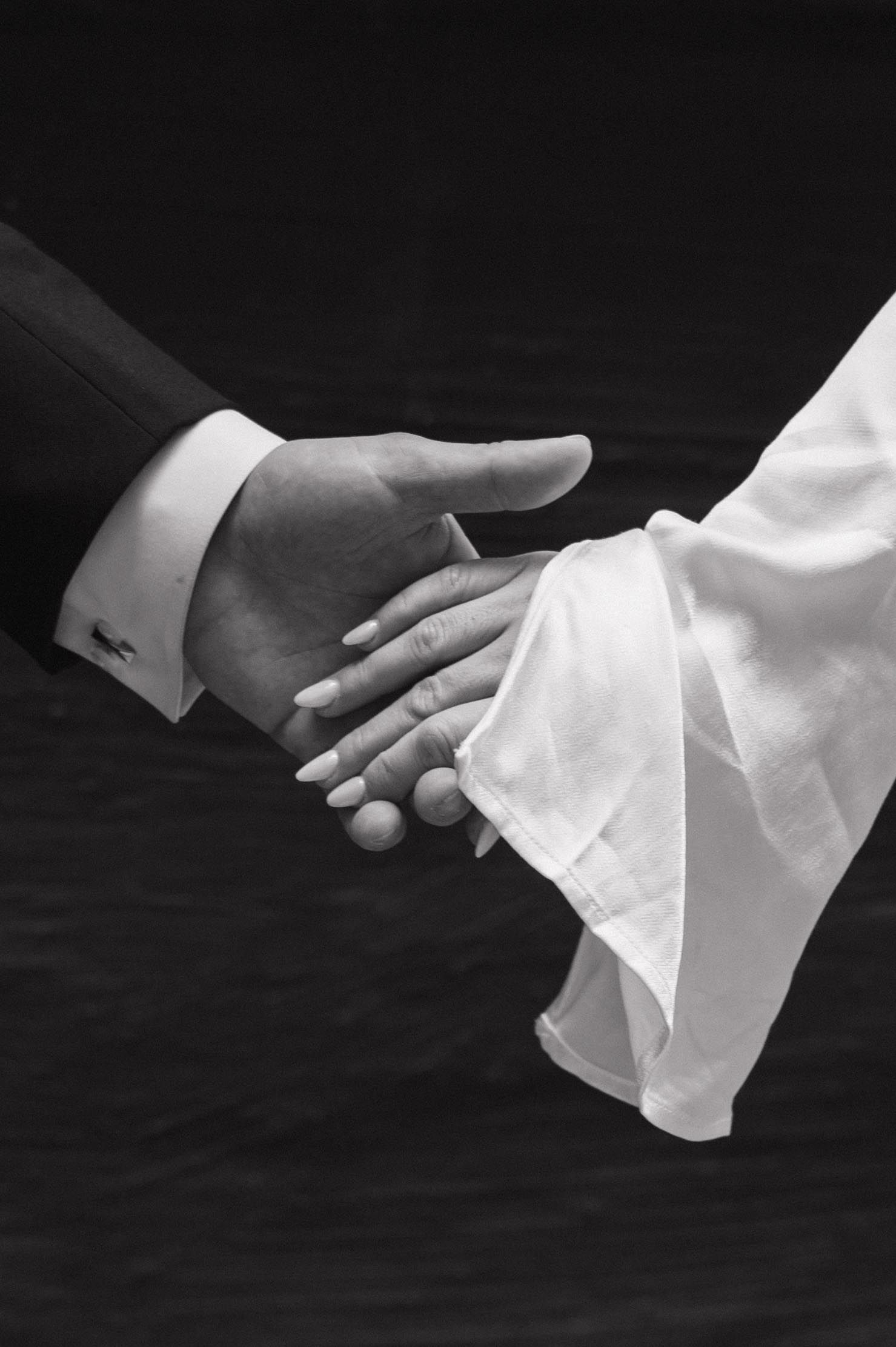 Black and white close-up of bride and groom holding hands, showcasing elegant wedding attire details.
