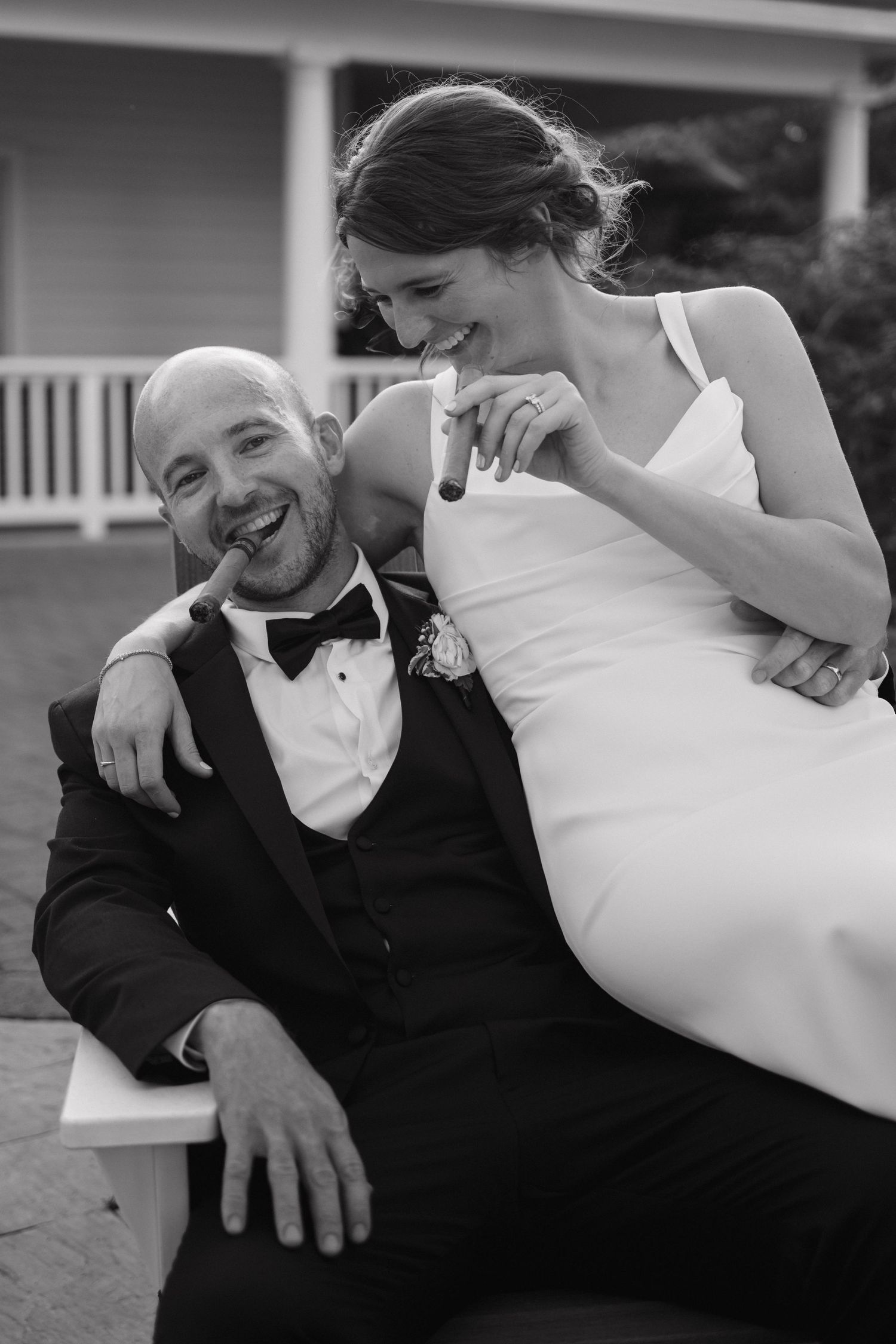 Black and white photo of laughing bride and groom smoking cigars together on their wedding day.