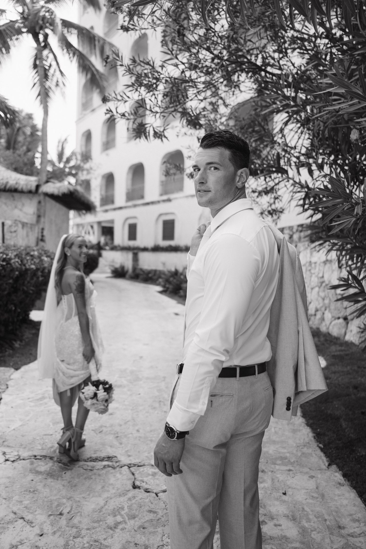 Groom in white shirt holds jacket while bride approaches in black and white wedding photo.