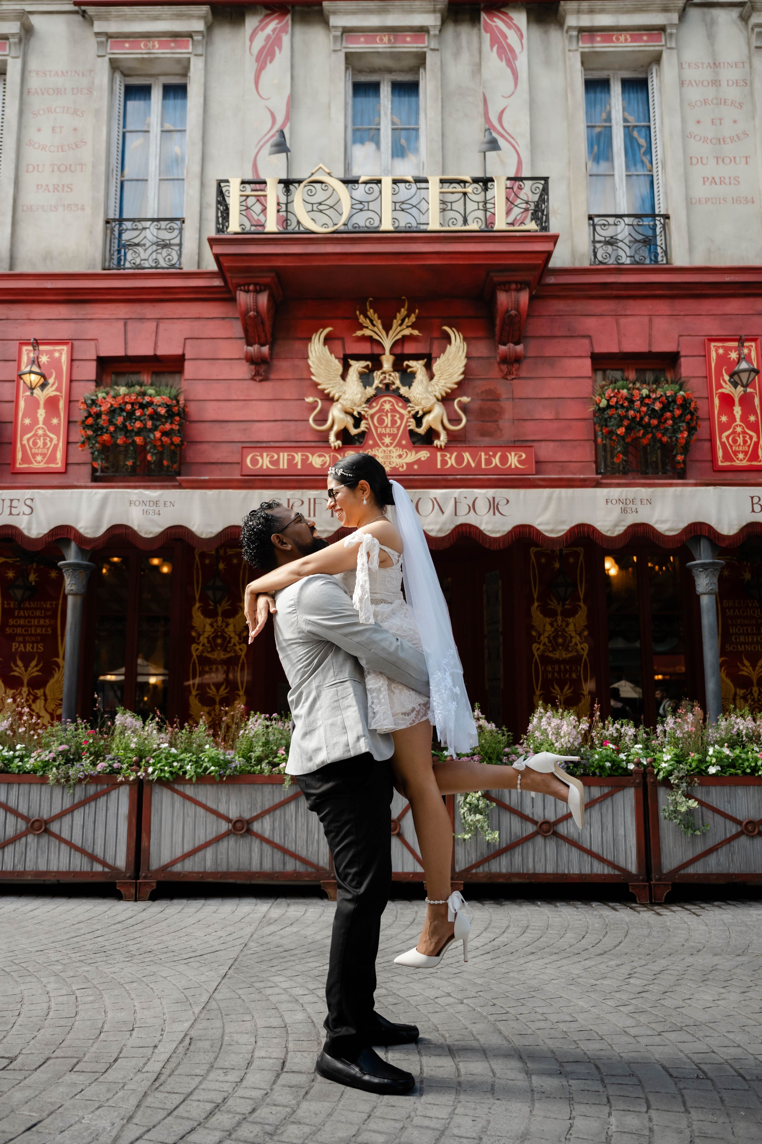 A couple shares a romantic embrace outside a red Parisian hotel, the groom lifting his bride in her wedding dress.